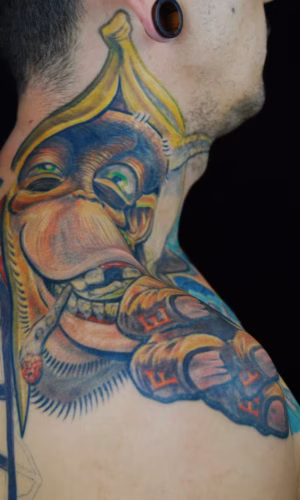 Kreatives Affen-Tattoo auf dem oberen Trapezius, anatomische Muskelform als Gesicht, Affe mit Bananen-Hut und Schriftzug ‚Affe‘ auf der Hand, Artist: Raul, München