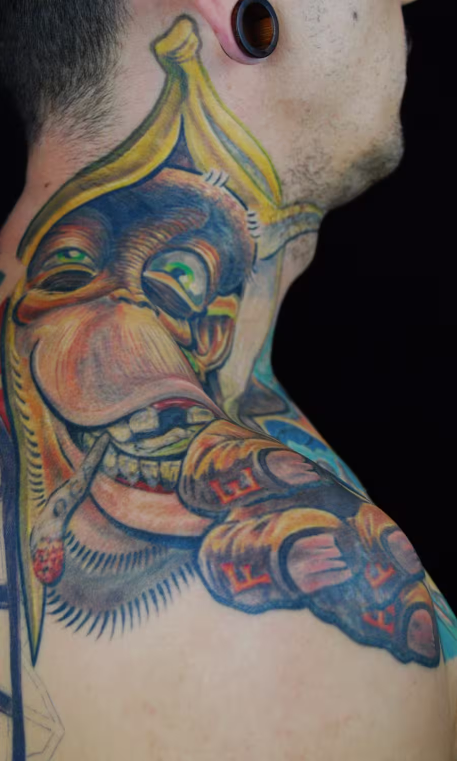 Kreatives Affen-Tattoo auf dem oberen Trapezius, anatomische Muskelform als Gesicht, Affe mit Bananen-Hut und Schriftzug ‚Affe‘ auf der Hand, Artist: Raul, München