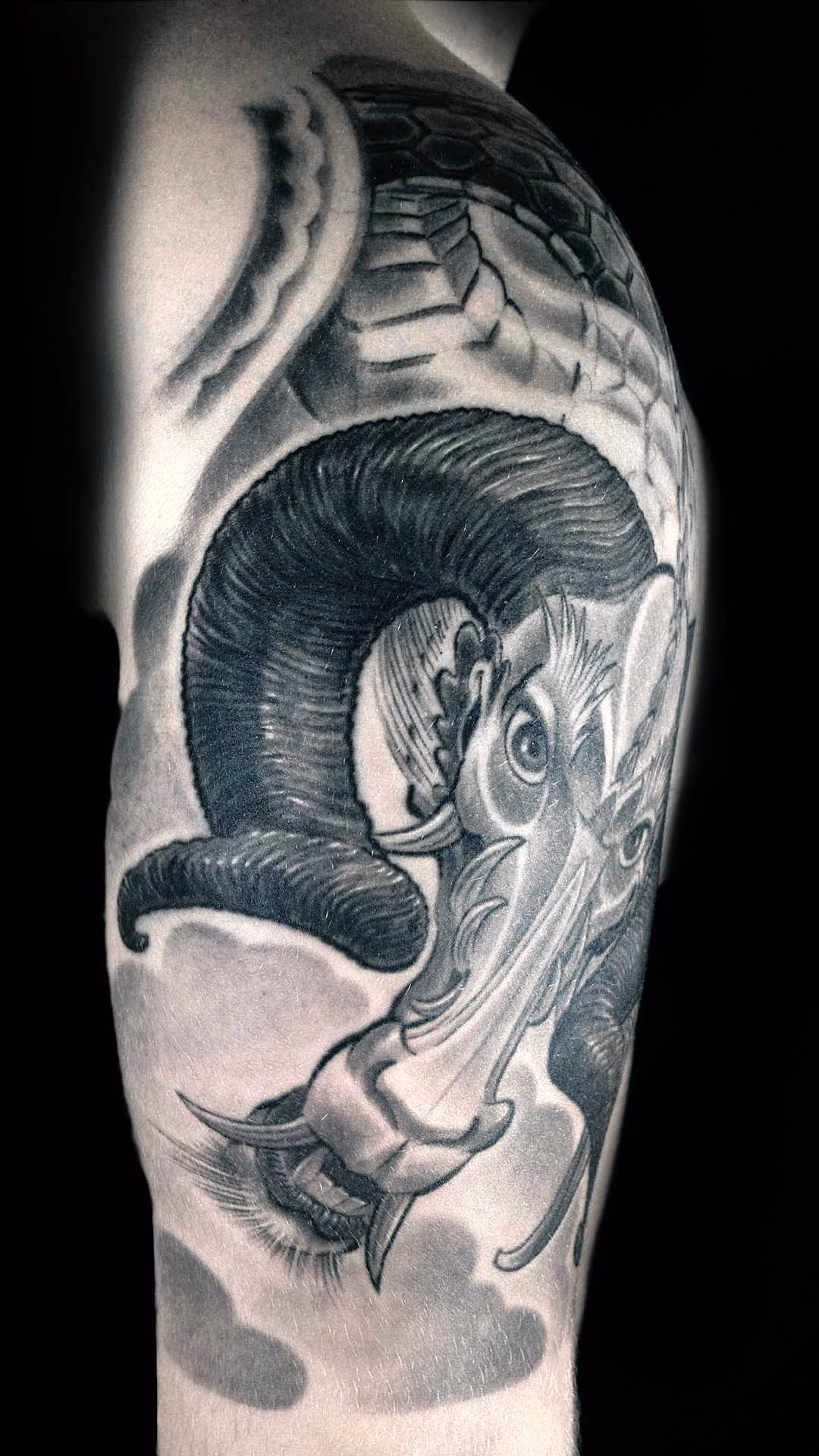 Widderdrache Japanisches Widder-Drachen Tattoo auf dem Oberarm. Detailreiches Black-and-Grey Custom Design mit eingerollten Hörnern vor einem Hintergrund aus weich schattierten Windbars und Wolken