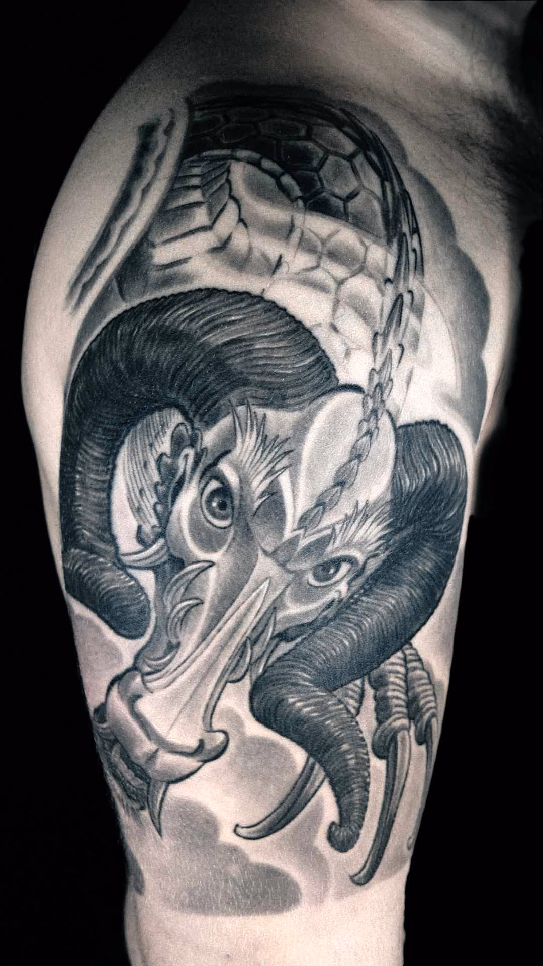 Widderdrache Großflächiges Black-and-Grey Tattoo auf dem männlichen Oberarm, lateral und leicht frontal. Es zeigt einen detaillierten japanischen Drachenkopf mit wuchtigen, eingerollten Widderhörnern und einer weichen Schnauze. Der Hintergrund ist mit sanften Schattierungen von traditionellen asiatischen Windbars und Wolken gestaltet