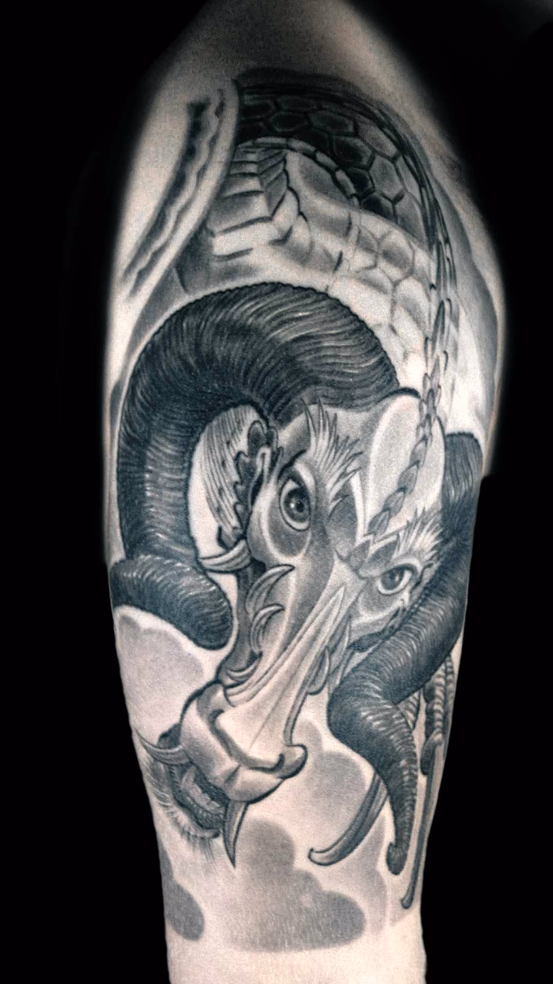 Widderdrache Oberarm Tattoo im Irezumi-Stil: Ein japanischer Drache mit individuellen Widderhörnern. Das Motiv nutzt die Anatomie des Deltamuskels perfekt aus. Weiche Schattierungen im Gesicht kontrastieren mit den dunklen Hörnern und den dynamischen Wolkenformationen im Hintergrund