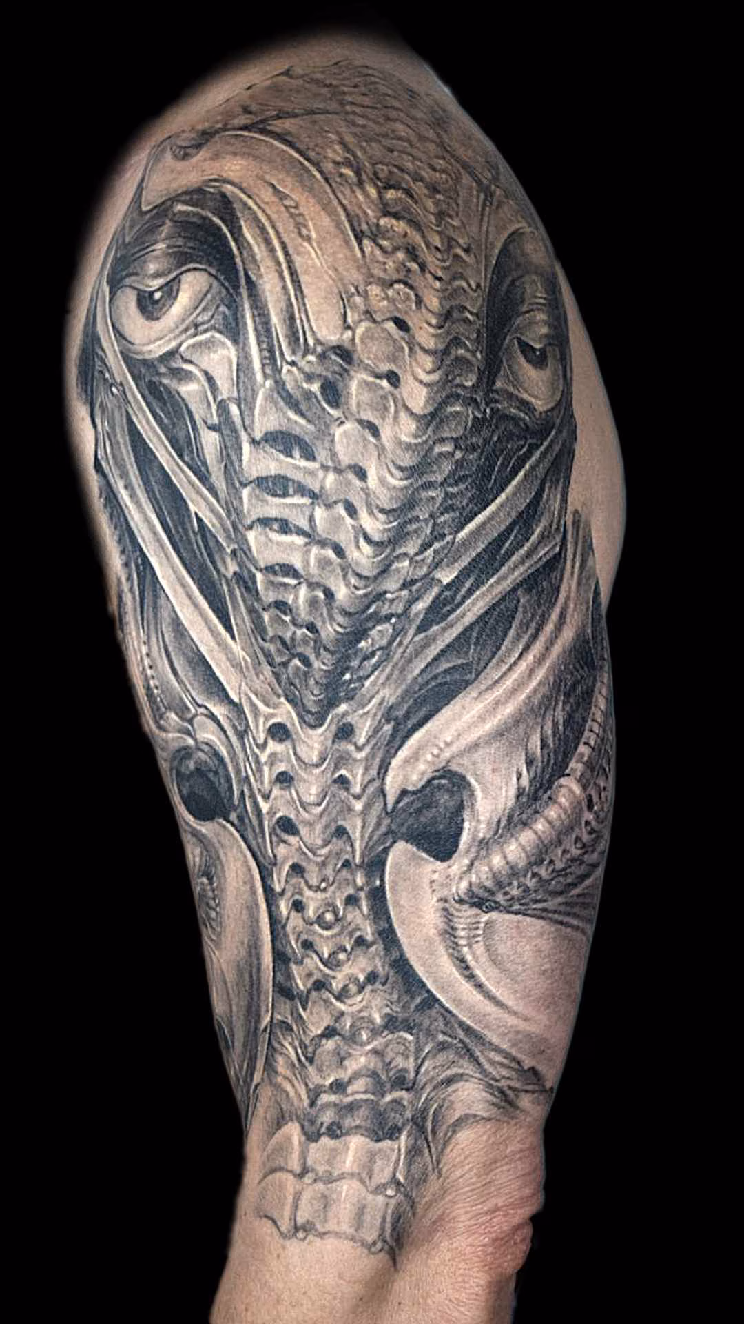 Template 1920 1080 mit Filtern Seitliche Perspektive des Custom Biomechanik-Tattoos auf der Tuberositas deltoidea. Die dunklen Schattierungen unterhalb des Deltamuskels betonen die Dreidimensionalität der insektenhaften Wirbelsäulen-Elemente und Knochenstrukturen
